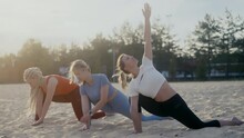 Three Athletic Beautiful Girls Doing Yoga Exercises On A Sandy Beach At Dawn On A Summer Morning. The Soft Light Of The Sun In The Background Fills The Scene. The Wind Shakes Hair In Slow Motion