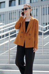 Poster - Young smiling woman in sunglasses and stylish casualwear talking on mobile phone while moving downstairs in urban environment