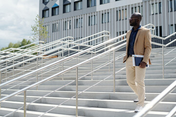 Wall Mural - Young serious African American male solopreneur with financial documents and tablet in hand walking downstairs against building