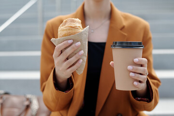 Hands of young modern businesswoman holding fresh croissant and cup of takeaway coffee while sitting in front of camera at lunch break