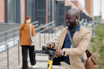 Poster - Young smiling African American businessman choosing settings on scooter terminal before riding home after work while standing in front of camera