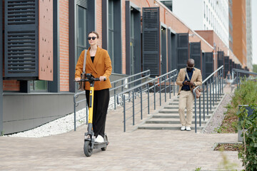 Wall Mural - Young active businesswoman in eyeglasses and quiet luxury apparel riding electric scooter and moving along modern building of office center