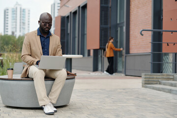 Wall Mural - Serious young male solopreneur in earphones typing and looking at screen of mobile device while sitting in urban environment and using laptop