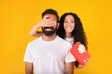 Wall Mural - Middle eastern woman closing man's eyes holding present, yellow background