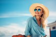 A stylish, smiling blond woman, suitcase in hand and dressed in trendy jeans, chic blouse, hat, and sunglasses, exudes the spirit of summer vacation and fashion