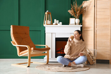 Poster - Young woman with cup of coffee near fireplace at home