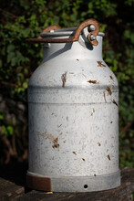 Close-up Of An Old Metal Milk Can. The Jug Is Outdoors In Bavaria And Is Partially Soiled With Manure. In The Background A Green Hedge. The Lid Is Closed.