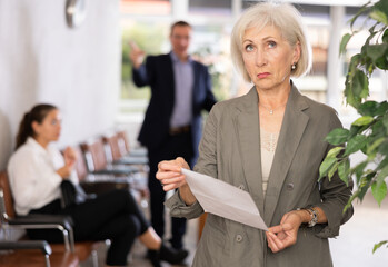 Wall Mural - Upset elderly woman in business suit posing with documents in her hands at reception..