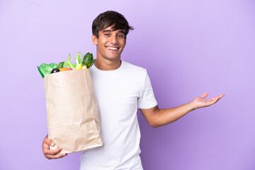 Wall Mural - Young man holding a grocery shopping bag isolated on purple background extending hands to the side for inviting to come