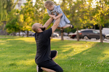 Wall Mural - young father throwing his daughter in the air on a summer day in park, happy moments in life