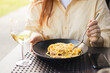 Caucasian young woman eating pasta carbonara and drinking white wine. Concept of italian lifestyle. Person eating dish from black plate outdoors
