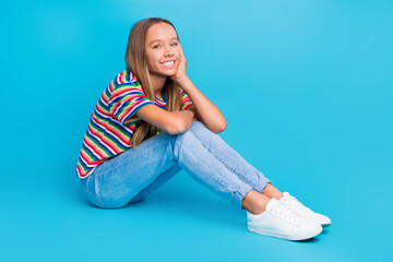 Sticker - Full size photo of optimistic cute teen girl wear colorful t-shirt pants sit on floor hand on cheekbone isolated on blue color background