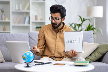 A young Indian male history and geography teacher teaches from home online via video link from a laptop. Shows the notebook to the camera, explains the lesson.