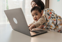 A Little Boy Asian Baby Is Disturbing His Mother While She Is Working At Home. Asian Mother Carrying Her Son Baby While Working On Computer Laptop At Home. Long Distance Working From Home Concept.