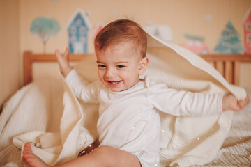 Wall Mural - A little baby in a white bodysuit sitting on a bed playing with a blanket and smiling