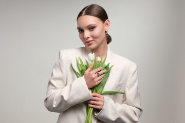 Wall Mural - Close up portrait of a beautiful elegant woman with stylish jewelry on a white gray background. Girl in white blazer holding tulips