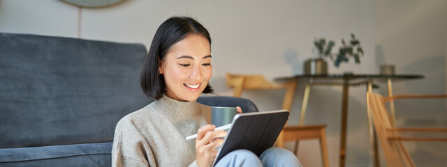 Wall Mural - Smiling korean woman sitting with tablet and pen, drawing and scatching her new project or design, working from home