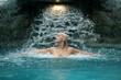 beautiful young sexy blonde nude woman, profile portrait stretches arms, enjoying the rippling splashing water of the waterfall in the Spa Wellness pool, feeling the falling water