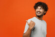 © ViDi Studio - Side view young cheerful happy Indian man he wear t-shirt casual clothes point thumb finger aside indicate on workspace area copy space mock up isolated on orange red color background studio portrait