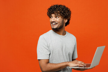 Side view young smiling happy Indian man wears t-shirt casual clothes hold use work on laptop pc computer look aside on area isolated on orange red color background studio portrait. Lifestyle concept.