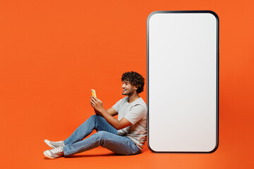 Wall Mural - Full body side view young Indian man wears t-shirt casual clothes sits near big huge blank screen mobile cell phone with workspace mockup area use smartphone isolated on orange red color background.