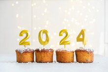 Golden Candles With 2024 Number On Top Of Cupcakes With Christmas Lights On Background. Merry Christmas And Happy New Year Celebration Mood. Happy Holidays. Festive Pastry Dessert.