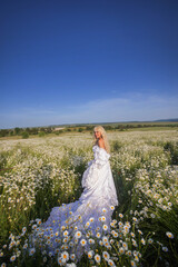 Wall Mural - Girl in a white dress and a crown with a bouquet in a chamomile field at sunset, view from the back, a princess on a walk