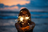 Fototapeta Tęcza - Young woman holding sparkler celebrating new years eve on the beach