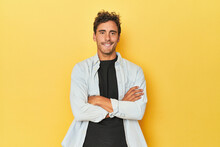 Young Latino man posing on yellow background who feels confident, crossing arms with determination.