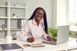 Portrait of friendly dark-skinned female financial advisor working with company accounting data. Woman is sitting at her workplace in front of laptop and calculator and is smiling at camera.