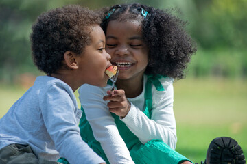 Happy boy and girl, African older sister feed her brother a piece of apple, family activities in summer