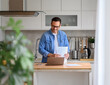 Smiling male financial adviser holding documents and working over laptop on kitchen counter at home