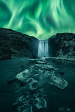 Aurora Borealis Dancing Over The Frozen Skógafoss Waterfall In Winter. A Man With A Lantern Is Watching Dance Across Night Skies The Aurora Polaris Next To The Famous Skógafoss Waterfall, Iceland
