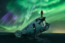 A Man With A Lantern Is Watching The Northern Lights (Aurora Borealis) Over A Douglas DC-3 Plane Wreckage. Cold Night, With Starry Sky And Polar Lights Over Abandoned Plane. Solheimasandur, Iceland