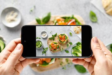 Poster - Female hands taking photo with smartphone of a healthy sandwiches with salted salmon, cream cheese, green romaine lettuce and fresh cucumber for breakfast to share on social media, top down view