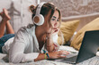© Daniel - Close up of one young pretty woman using laptop indoor at home lying on the bed wearing headphones listening music. Millennial teenager having fun online studying. .