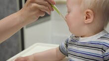 Mother feeding son with baby food from spoon. Child eats porridge sitting on high chair. Mom feeds baby fruit puree in kitchen at home.