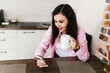 © Mila - A young girl with black hair in a pink shirt sits at a table with a phone and a white mug in a cozy room with brown brick. Woman working, talking on the phone, drinking coffee in a modern cafe.Morning