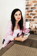 © Mila - A young girl with black hair in a pink shirt sits at a table with a phone and a white mug in a cozy room with brown brick. Woman working, talking on the phone, drinking coffee in a modern cafe.Morning