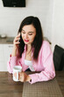 © Mila - A young girl with black hair in a pink shirt sits at a table with a phone and a white mug in a cozy room with brown brick. Woman working, talking on the phone, drinking coffee in a modern cafe.Morning