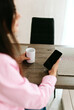 © Mila - A young girl with black long hair in a pink shirt and white top sits at a table with a phone and a white mug in a cozy room. A white mug and a phone in the hands of a girl. Morning business woman