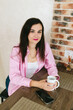 © Mila - A young girl with black hair in a pink shirt sits at a table with a phone and a white mug in a cozy room with brown brick. Woman working, talking on the phone, drinking coffee in a modern cafe.Morning