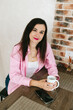© Mila - A young girl with black hair in a pink shirt sits at a table with a phone and a white mug in a cozy room with brown brick. Woman working, talking on the phone, drinking coffee in a modern cafe.Morning