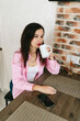© Mila - A young girl with black hair in a pink shirt sits at a table with a phone and a white mug in a cozy room with brown brick. Woman working, talking on the phone, drinking coffee in a modern cafe.Morning