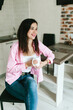 © Mila - A young girl with black hair in a pink shirt sits at a table with a phone and a white mug in a cozy room with brown brick. Woman working, talking on the phone, drinking coffee in a modern cafe.Morning