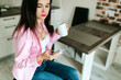 © Mila - A young girl with black long hair in a pink shirt and white top sits at a table with a phone and a white mug in a cozy room. A white mug and a phone in the hands of a girl. Morning business woman