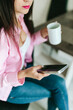 © Mila - A young girl with black long hair in a pink shirt and white top sits at a table with a phone and a white mug in a cozy room. A white mug and a phone in the hands of a girl. Morning business woman