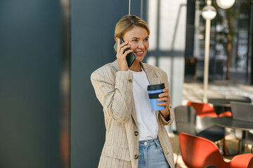 Smiling business woman holding coffee and talking phone standing on modern office background 