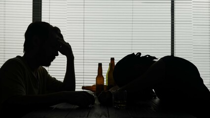 Two men are sitting at a table in a cafe. They sit next to empty bottles, they drank a lot. One man has fallen asleep at the table, the second man sits and is sad. Demonstrating alcoholism