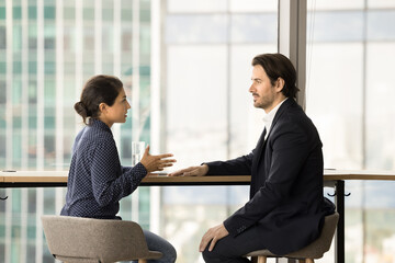 Serious confident diverse colleagues discussing project strategy, cooperation in co-working space. Young female Indian employee talking to male boss, company business leader at large window